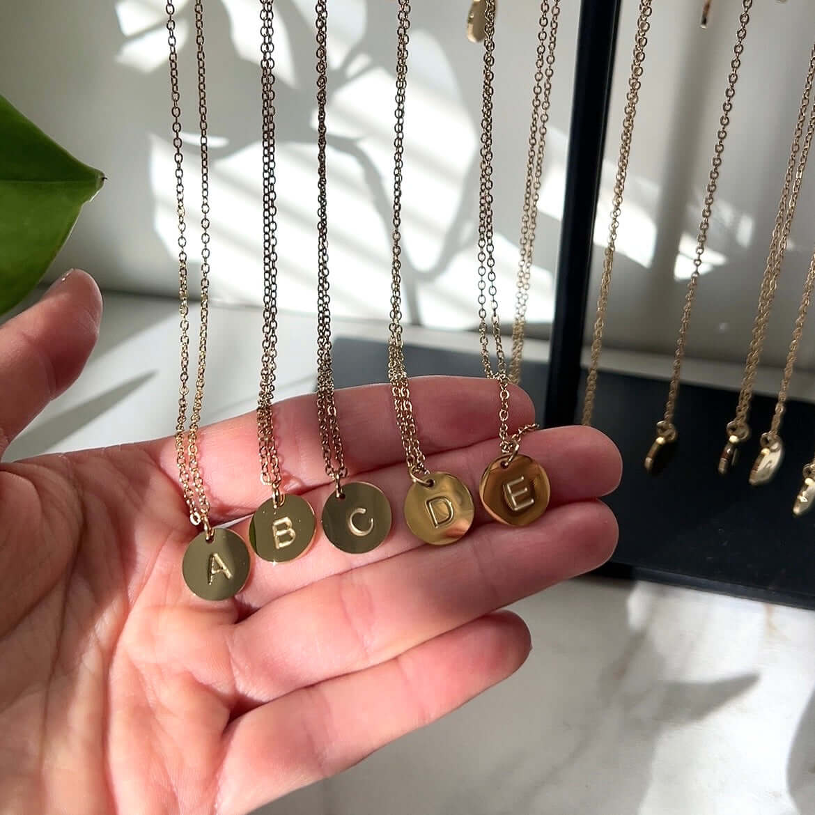 Käsi pitelee viittä korutelineestä roikkuvaa kaulakorua, joissa kirjaimet A, B, C, D ja E.