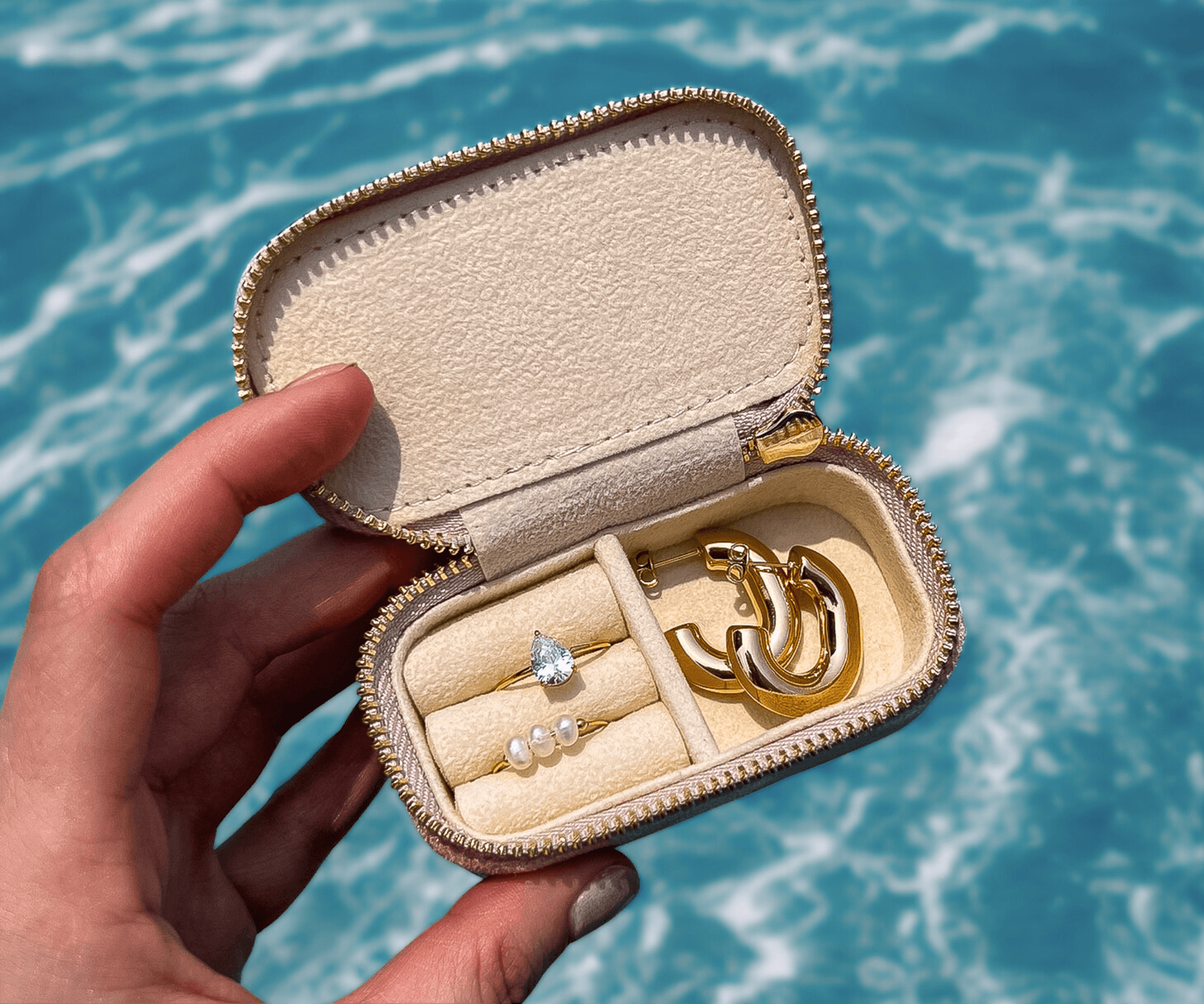 Käsi pitelee pientä vaaleanpunaista korurasiaa, jossa kaksi kultaista sormusta ja korvakorut. Taustalla sinistä vettä.